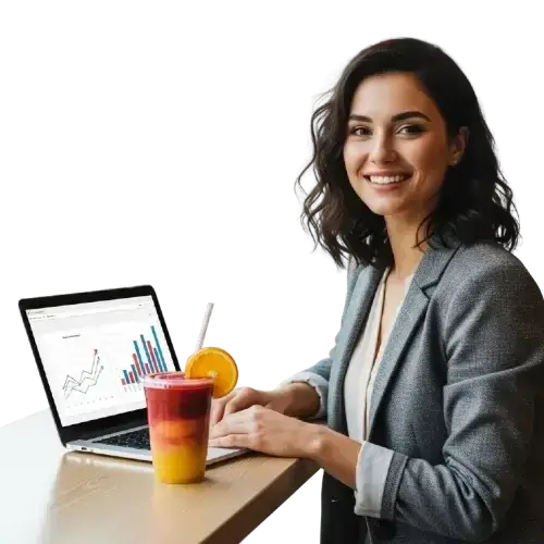 Jovem empreendedora sorrindo ao lado de um laptop com gráficos financeiros de crescimento e um copo de smoothie colorido em camadas com rodela de laranja sobre a mesa.